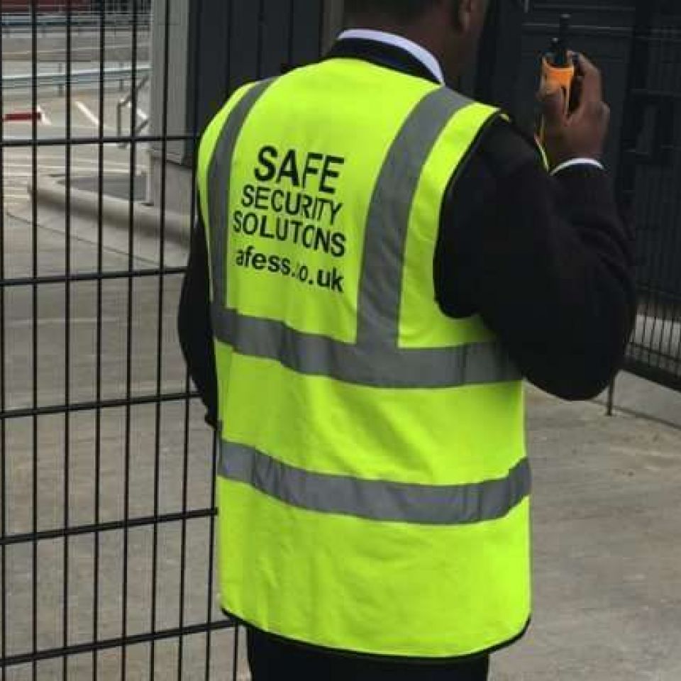 A Safe Security Solutions Staff Picture of a man in a yellow branded vest holding a walkie talkie.