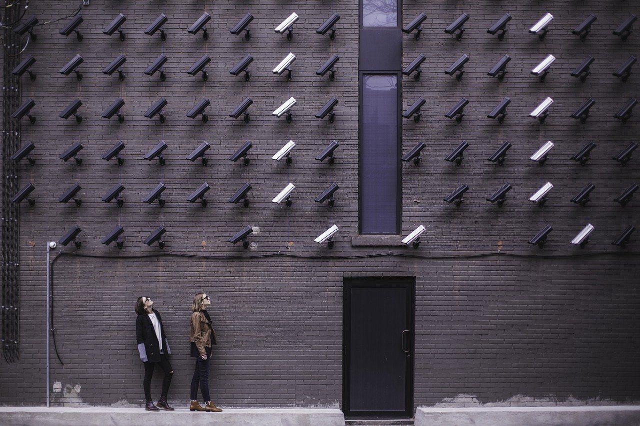 A wall with lots of CCTV cameras mounted. there are two women looking up at them